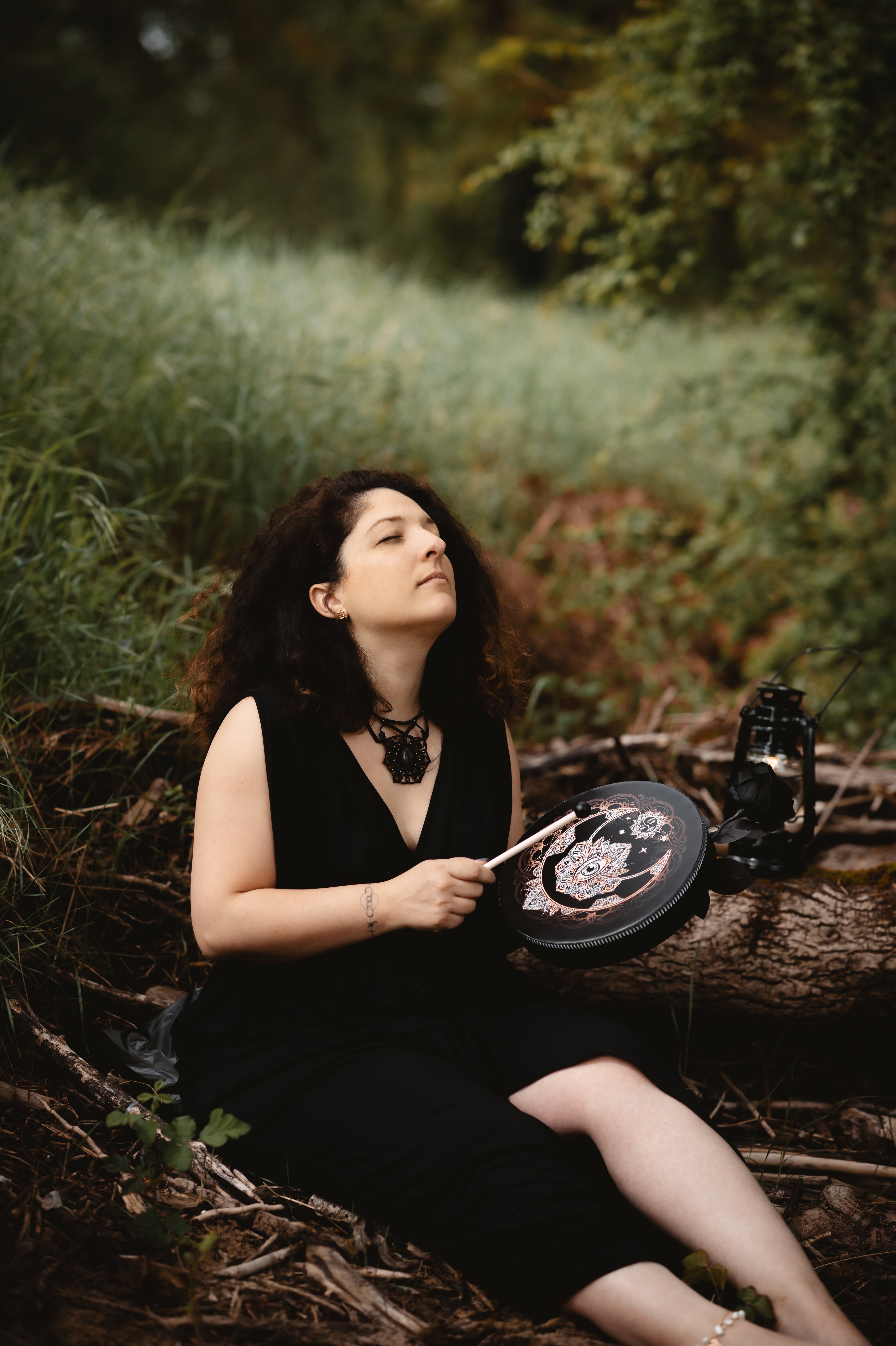Emilie, fondatrice, assise en extérieur dans un sous-bois, les yeux fermés, tenant un tambour orné de motifs spirituels. Vêtue de noir, elle joue avec une mailloche, entourée de végétation et de branches au sol.
